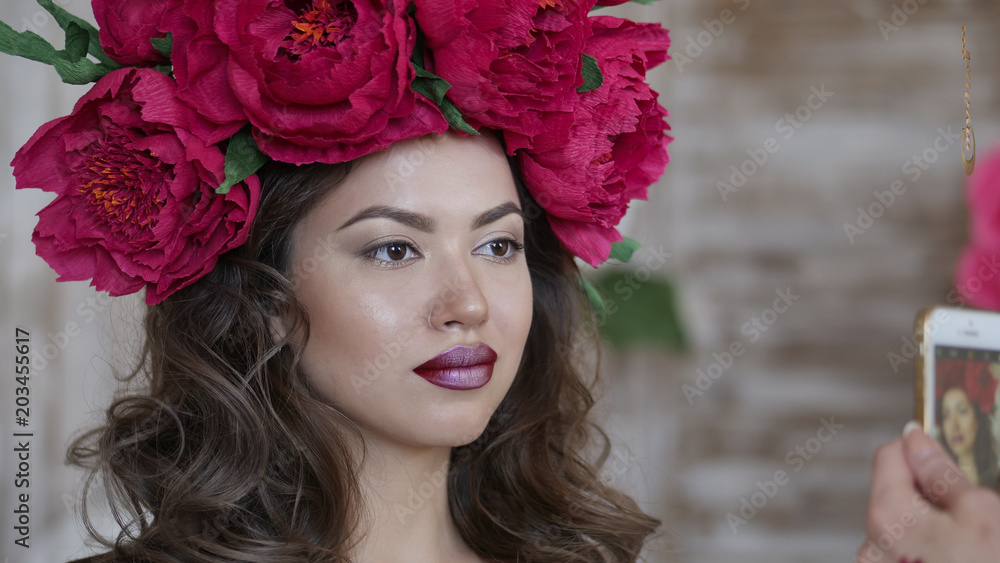 Beautiful model, with a wreath of scarlet peonies on his head. The model is photographed on the phone, a smartphone Beautiful model, with a wreath of scarlet peonies on his head. The model is photographed on the phone, a smartphone