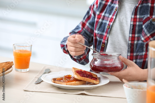 Cute little boy spreading jam onto tasty toasted bread at table