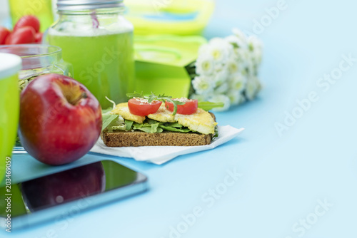 Healthy vegetarian Breakfast. Sandwich Grain bread with herbs and tofu, detox smoothie, apple, sunflower seeds, cherry tomatoes, fresh natural flowers, blue background, smartphone. Copy space for text