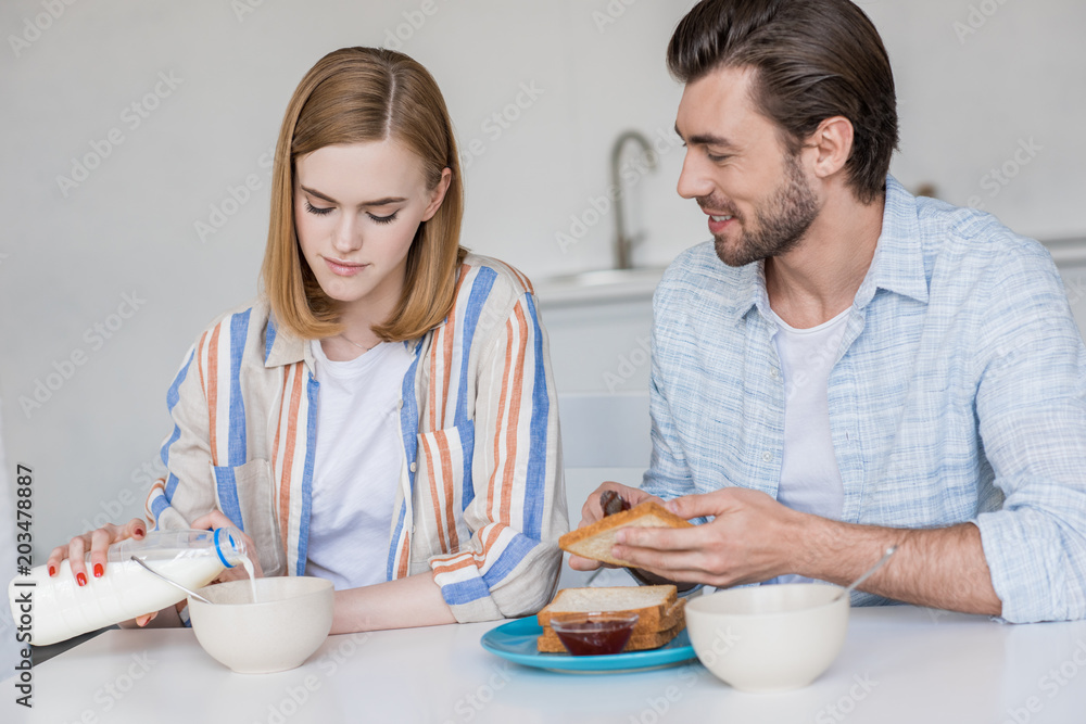 young woman pouring into bowl and boyfriend spreading jam on toast young woman pouring into bowl and boyfriend spreading jam on toast