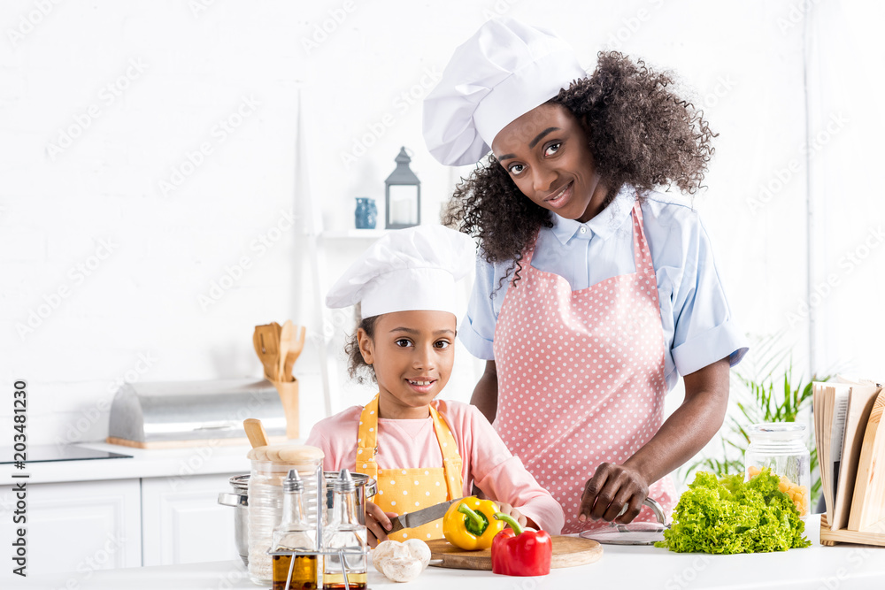 happy african american mother and daughter in chef hats cutting bell pepper on kitchen happy african american mother and daughter in chef hats cutting bell pepper on kitchen