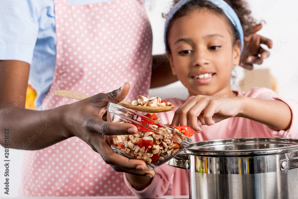 african american mother and daughter putting ingredientes in saucepan on kitchen african american mother and daughter putting ingredientes in saucepan on kitchen