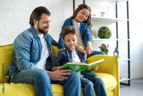 smiling father with two kids reading book together while sitting on couch at home