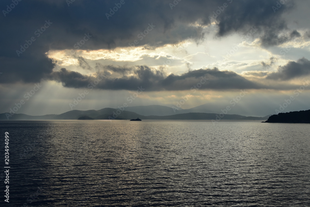 Early morning, dramatic sky, dark steel Ionian Sea waters. Peristeres lighthouse on a small rocky islet seen from Kassiopi, Corfu Island, Greece. Albanian coast on the horizon.