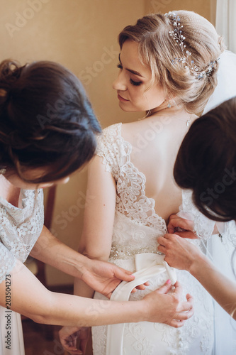 Beautiful bridesmaids help put on wedding dress on gorgeous blonde bride in luxury hotel room morning before the wedding