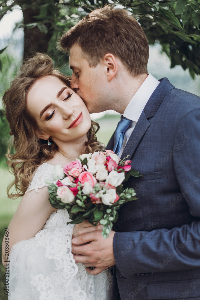Sensual groom kissing beautiful bride on the cheek, newlywed couple portrait, beautiful married couple posing outdoors Sensual groom kissing beautiful bride on the cheek, newlywed couple portrait, beautiful married couple posing outdoors