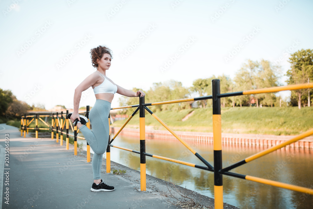 Fototapeta premium Beautiful sporty girl with curly hair is stretching her leg outdoors