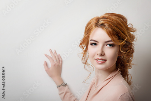 Red-haired girl with make-up leans her hand against a white wall and smiles.