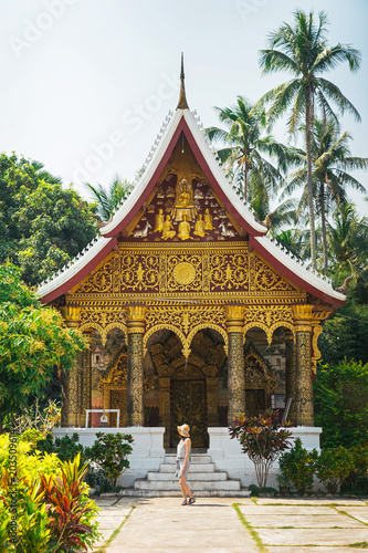 The tourist girl stands on the background of a beautiful Asian temple in Laos, Luang Prabang.Trip to Laos.Young woman walks among green tropical plants against the backdrop of a beautiful temple.