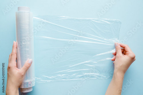 Woman's hand using a roll of transparent polyethylene food film for packing products on the pastel blue table. Empty place for text or logo.