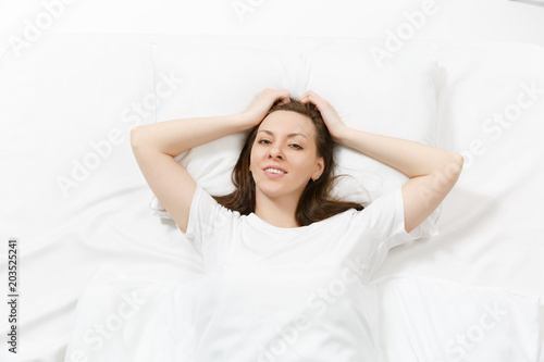 Top view of head of happy brunette young woman lying in bed with white sheet, pillow, blanket. Calm beautiful female spending time in room. Rest, relax, good mood concept. Copy space for advertisement