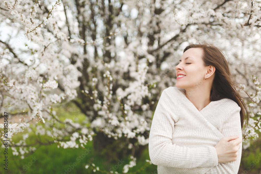 Young smiling relaxed tender beautiful woman with closed eyes in light casual clothes standing in city garden or park on blooming tree background. Spring nature, flowers. Lifestyle, leisure concept. Young smiling relaxed tender beautiful woman with closed eyes in light casual clothes standing in city garden or park on blooming tree background. Spring nature, flowers. Lifestyle, leisure concept.