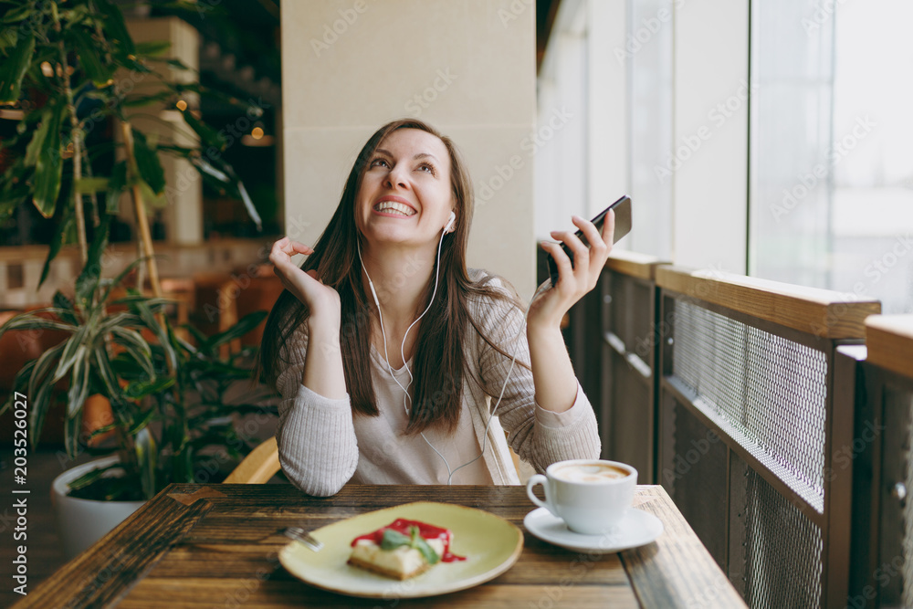 Young woman sitting in coffee shop at table with cup of cappuccino, cake, relax in restaurant during free time. Young female listen music in earphones on mobile phone, rest in cafe. Lifestyle concept. Young woman sitting in coffee shop at table with cup of cappuccino, cake, relax in restaurant during free time. Young female listen music in earphones on mobile phone, rest in cafe. Lifestyle concept.