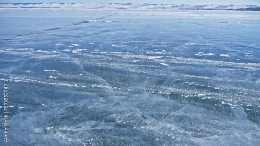 Snow is flying over surface of ice. Snowflakes fly on ice of Lake Baikal. Ice is very beautiful with unusual unique cracks. Background view mountain landscape. Snow sparkles and glows in red. Shooting Snow is flying over surface of ice. Snowflakes fly on ice of Lake Baikal. Ice is very beautiful with unusual unique cracks. Background view mountain landscape. Snow sparkles and glows in red. Shooting