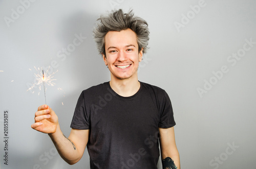 Young guy in black t-shirt holds sparkler and smiles, on gray background. photo