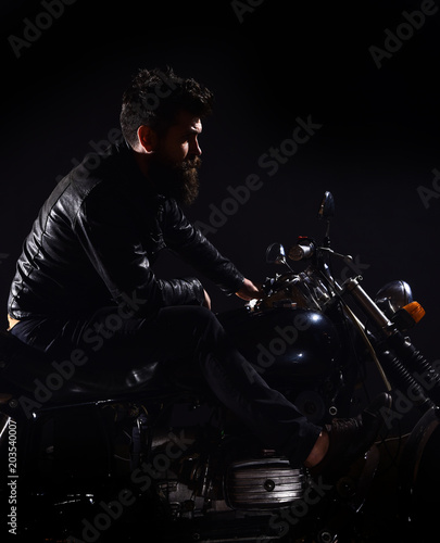 Macho, brutal biker in leather jacket riding motorcycle at night time, copy space. Man with beard, biker in leather jacket sitting on motor bike in darkness, black background. Bikers leisure concept.