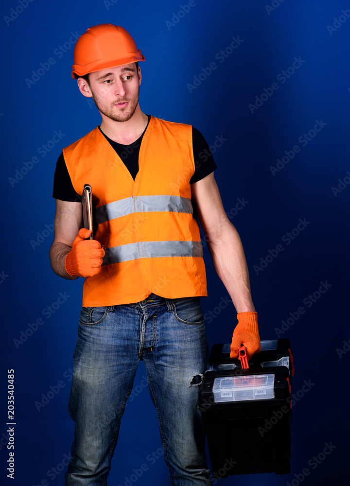 Man in helmet, hard hat holds toolbox and folder with documents, blue background. Worker, repairer, builder on dreamy face carries case with tools. Repair service concept. Repairman ready to work. Man in helmet, hard hat holds toolbox and folder with documents, blue background. Worker, repairer, builder on dreamy face carries case with tools. Repair service concept. Repairman ready to work.
