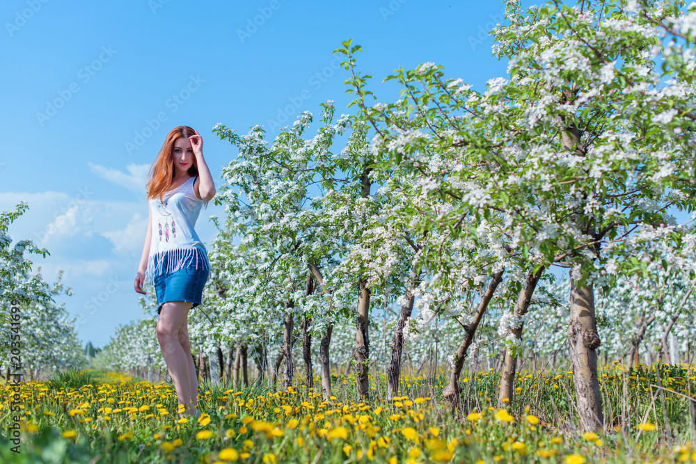 Fototapeta premium Beautiful girl in apple blossom garden on a spring day. Summer romantic image of stylish casual woman in t shirt and jeans skirt , trendy and fashionable accessories . positive mood