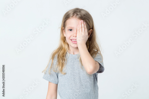 Little girl closed one eye with her hand, standing on a light background.