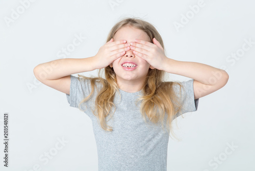 I do not see. Little girl covered her eyes with her hands, standing on a light background.