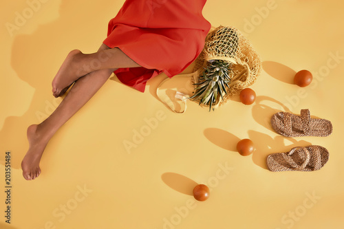 Legs of black woman in orange dress sitting on yellow background with oranges, pineapple in net bag and straw shoes on the floor photo