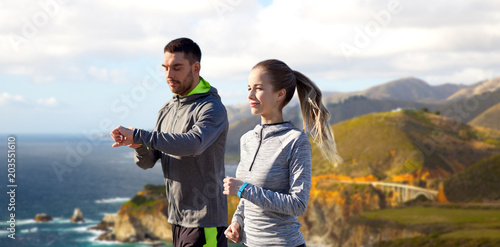 fitness, sport and healthy lifestyle concept - smiling couple with heart-rate watches running over bixby creek bridge on big sur coast of california background