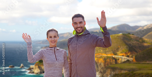 fitness, sport, and healthy lifestyle concept - smiling couple waving hand over bixby creek bridge on big sur coast of california background