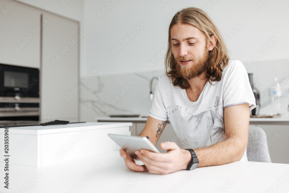 Portrait of young smiling man sitting and using digital tablet in kitchen at home isolated Portrait of young smiling man sitting and using digital tablet in kitchen at home isolated