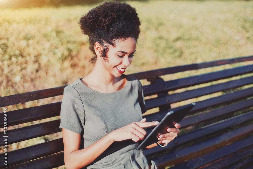 Young curly black smiling female with a digital pad on the bench in the park; charming cheerful Brazilian girl with Afro hair using tablet pc while sitting on the wooden bench on a summer day Young curly black smiling female with a digital pad on the bench in the park; charming cheerful Brazilian girl with Afro hair using tablet pc while sitting on the wooden bench on a summer day
