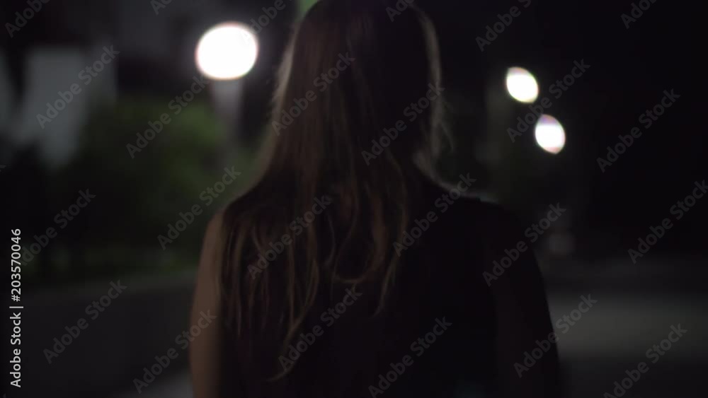 Back view of a young woman walking in desert poorly lighted street. She is scared to be alone outside at night