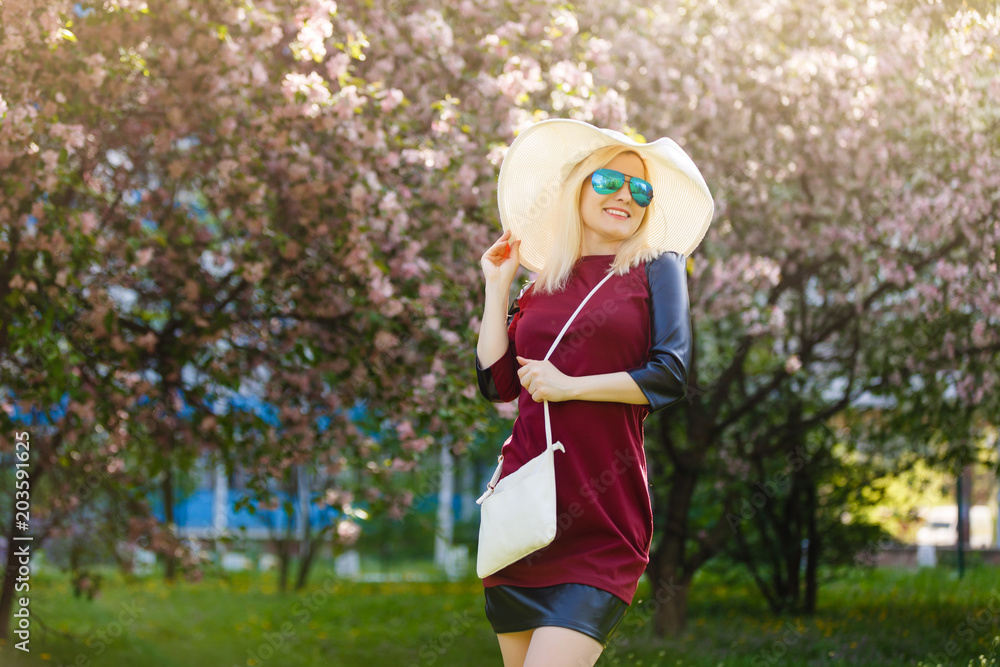 Fototapeta premium Summer fashion portrait of pretty blonde woman posing on amazing blooming tree background .Wearing sunglasses, casual hat , trendy blue silk kimono and velvet choker. Stylish handbag.