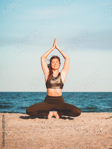 Young beautiful sporty woman in green clothing doing yoga asana on sea beach near water. Girl practicing exercises. Health concept.