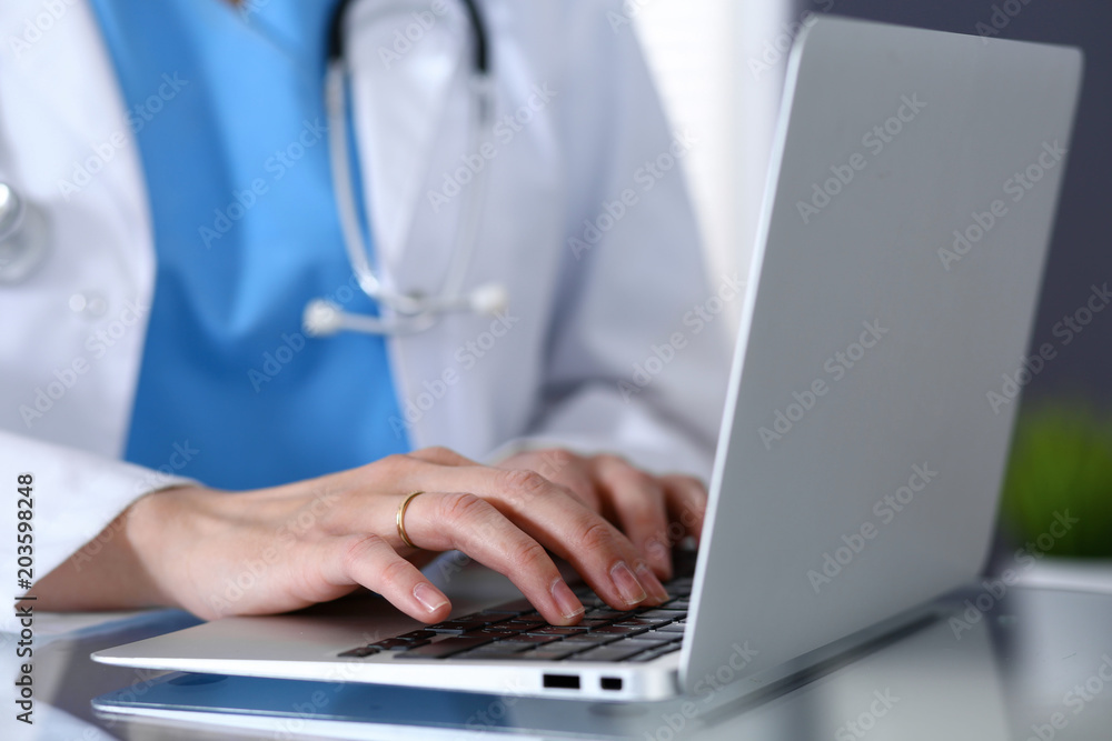Doctor typing on laptop computer while sitting at the glass desk in hospital office. Physician at work. Medicine and healthcare concept