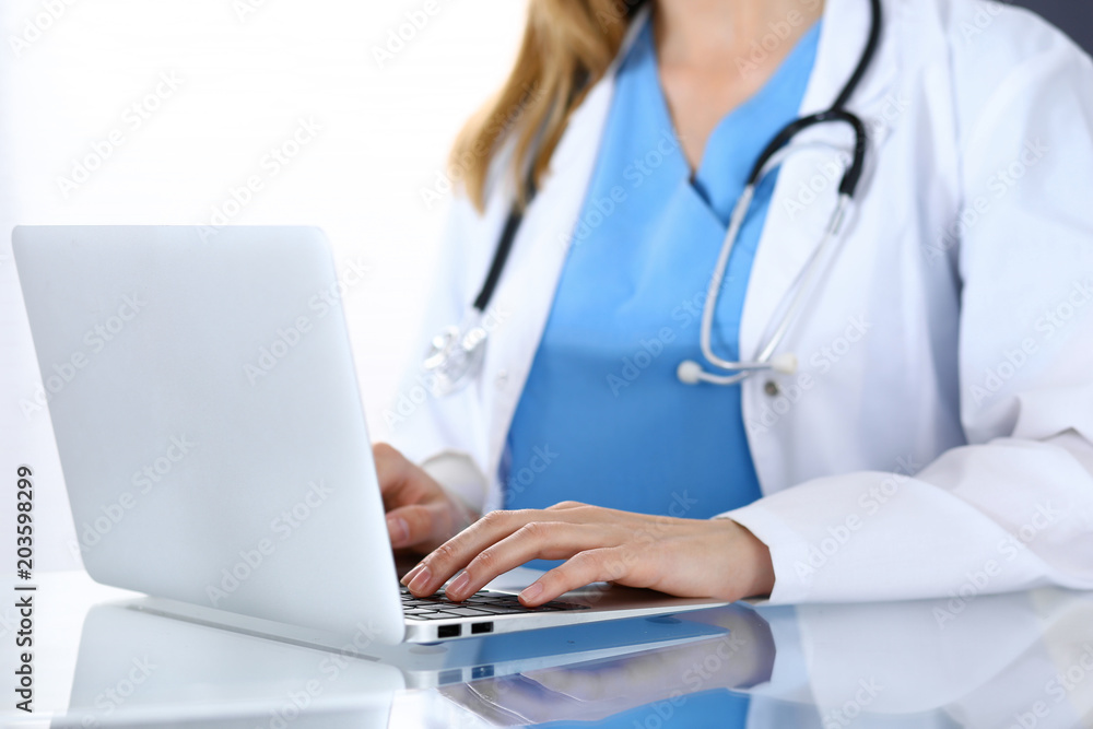 Doctor typing on laptop computer while sitting at the glass desk in hospital office. Physician at work. Medicine and healthcare concept Doctor typing on laptop computer while sitting at the glass desk in hospital office. Physician at work. Medicine and healthcare concept