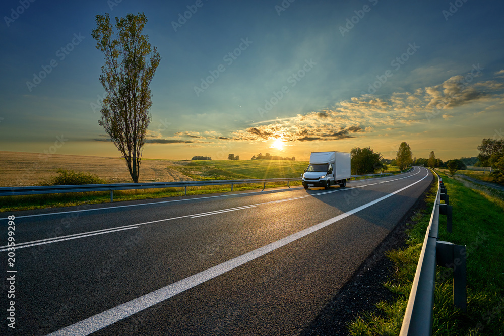White delivery van driving on asphalt road around farm fields in rural landscape at sunset