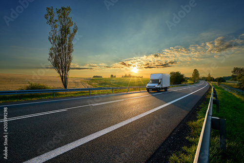 White delivery van driving on asphalt road around farm fields in rural landscape at sunset photo