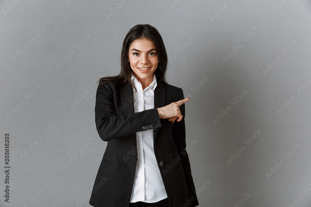 portrait of smiling businesswoman in suit pointing away against grey wall backdrop portrait of smiling businesswoman in suit pointing away against grey wall backdrop