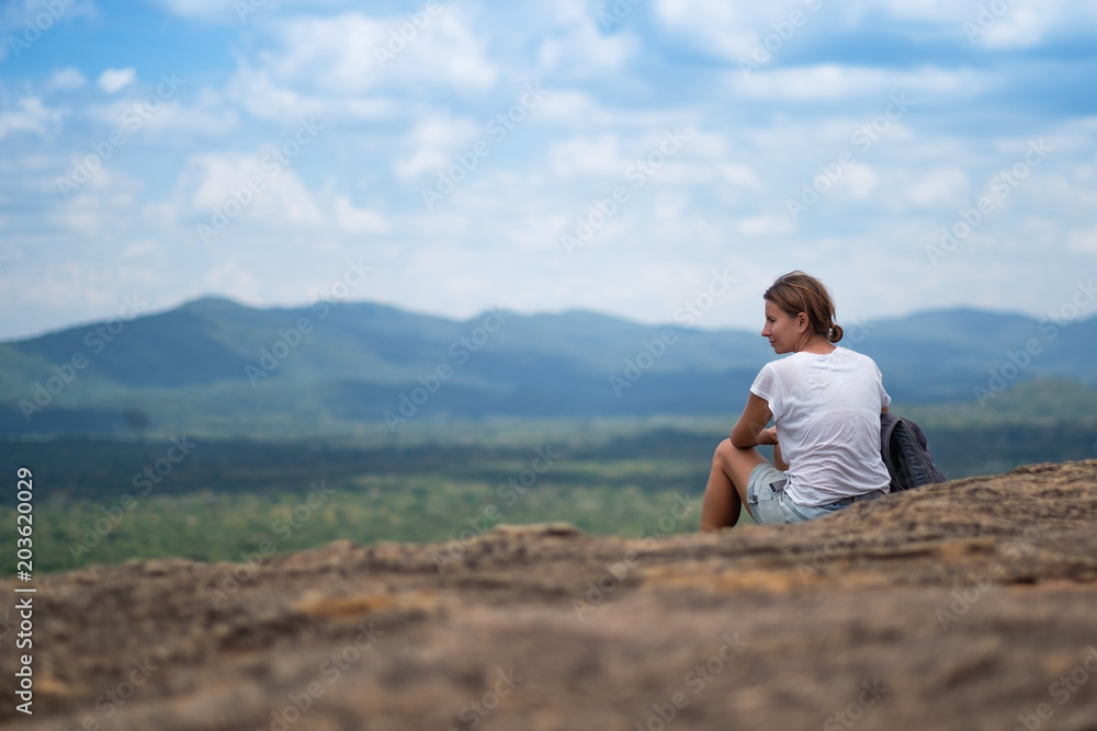 Young woman with backpack sitting on mountain and looking to a sky with clouds. Beautiful nature of Sri Lanka