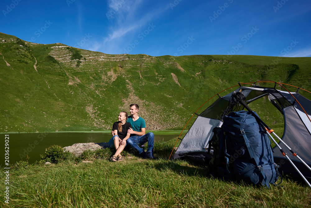Romantic couple sits on rock near tent, backpack and trekking sticks and look into the distance against background of beautiful landscape of green mountains and lake and their foothills under blue sky