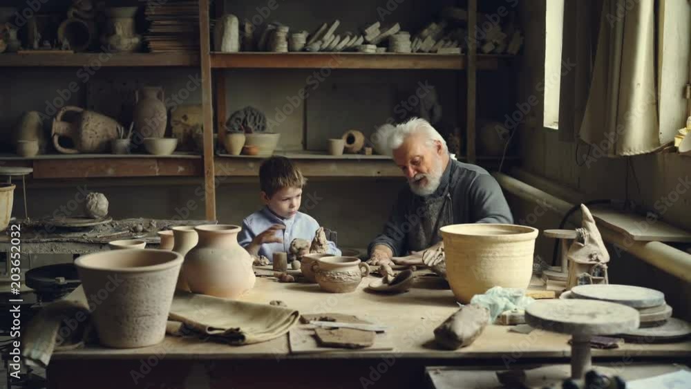 Experienced craftsman is teaching his grandchild pottery art and making clay rolls for ceramic figures while curious boy is playing with large piece of clay. Experienced craftsman is teaching his grandchild pottery art and making clay rolls for ceramic figures while curious boy is playing with large piece of clay.