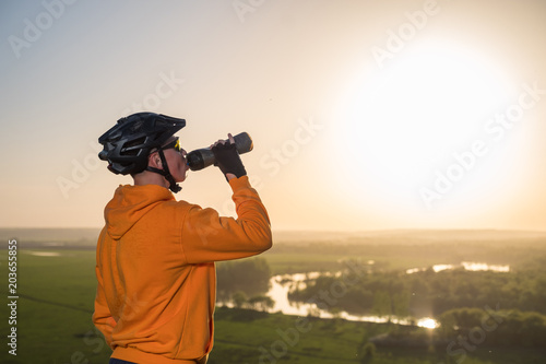 Cyclist at sunset in the mountains. A young man in a helmet and glasses drinks water from a bottle.