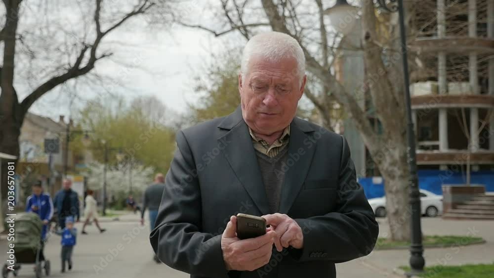 An imprressive closeup of a wise aged man in a jacket, who surfs the net and walks along a city street with modern houses in spring in slo-mo An imprressive closeup of a wise aged man in a jacket, who surfs the net and walks along a city street with modern houses in spring in slo-mo