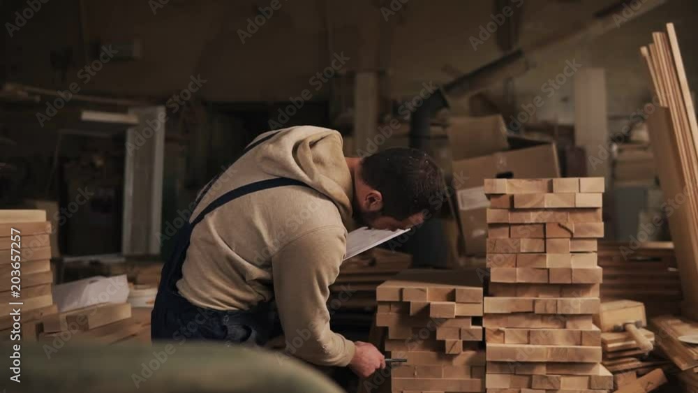 A man in a carpentry workshop with a tablet and a pen counts the number of the wood blocks. Jointer is in the working clothes. Front view A man in a carpentry workshop with a tablet and a pen counts the number of the wood blocks. Jointer is in the working clothes. Front view