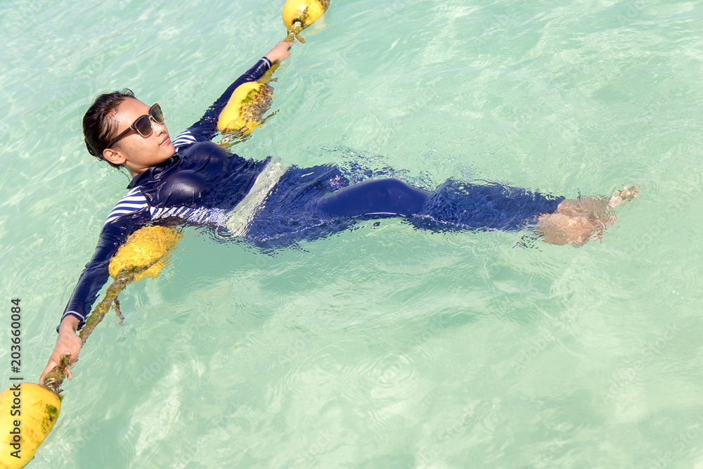 Woman in a full-body swimsuit lying on yellow buoys in the clear sea. Girl in blue burkini relaxes in a turquoise sea.