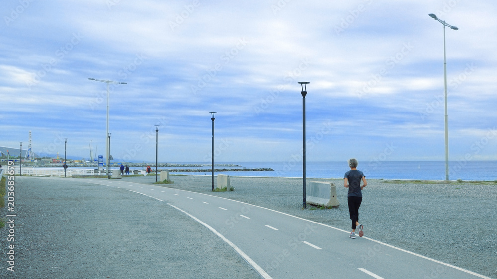Senior woman running in a promenade against a blue cloudy sky. Active ageing concept and empty copy space for Editor's text.