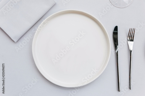 Top view of white empty plate on white tablecloth with knife, fork, napkin and wine glass.
