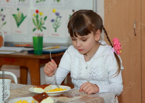 A beautiful little girl is having Breakfast in the kindergarten. She eats casserole and corn.