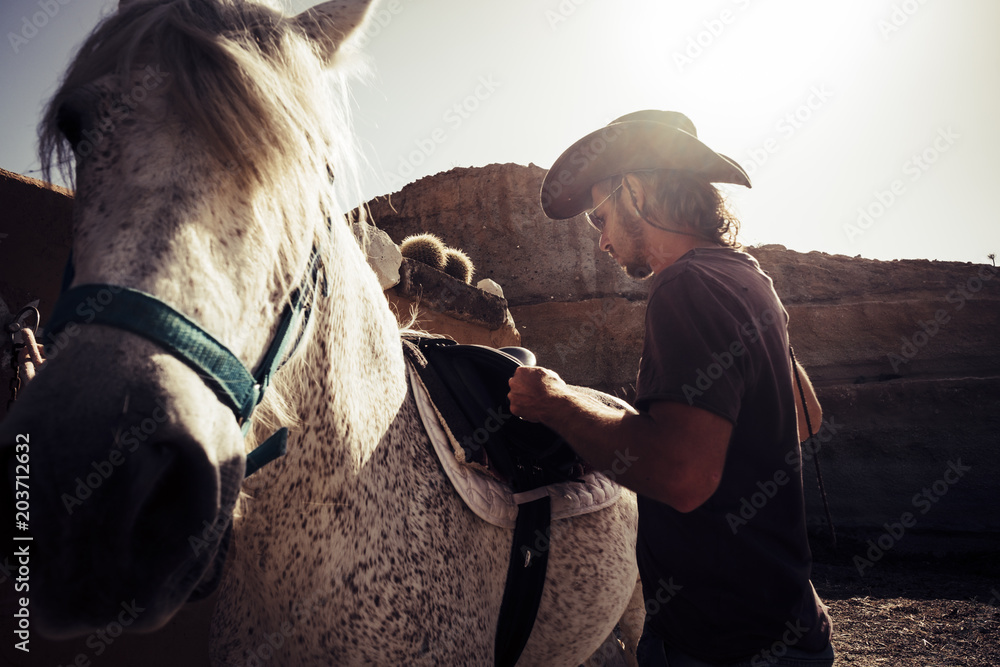 Obraz premium beautiful cowboy scene with mountains on the background and backlight from the sunset for beautiful young man blonde long hair and best friend horse together ready to live leisure outdoor activity
