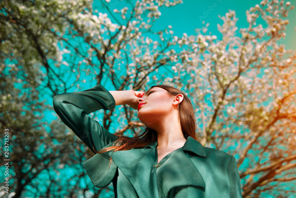 Fototapeta premium Beauty fashion model girl posing over blooming trees, enjoying nature in spring apple orchard. Beautiful brunette young woman over sky in Garden with blooming apple trees. blossom flowers outdoors