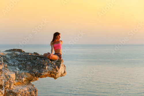 Meditation on the beach. A woman sits on a cliff above the ocean and does yoga. Femininity. Force. Soft shades at sunset. Beautiful scenery. You're on top of the world.